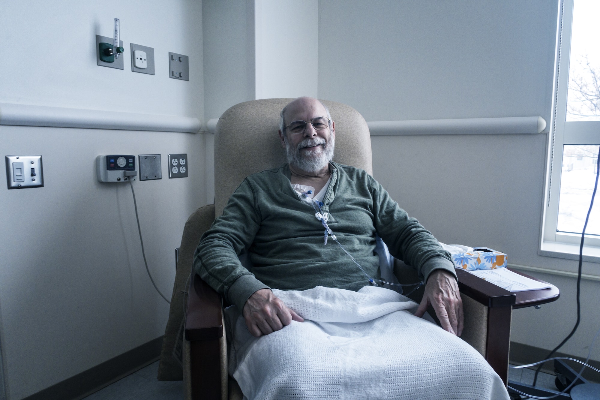 A senior adult man outpatient cancer patient - alert, but tired and bored - is sitting resting comfortably while chemotherapy IV drip medicine is administered by a tangled array of medical equipment through an intravenous chemo access port temporarily embedded into his upper chest. The plastic tubes, clamps, connectors, caps and off-camera drip bags are attached only during each two to three hour on-site session at this medical hospital clinic. The embedded access port stays in place throughout his three month bi-weekly chemotherapy regimen.